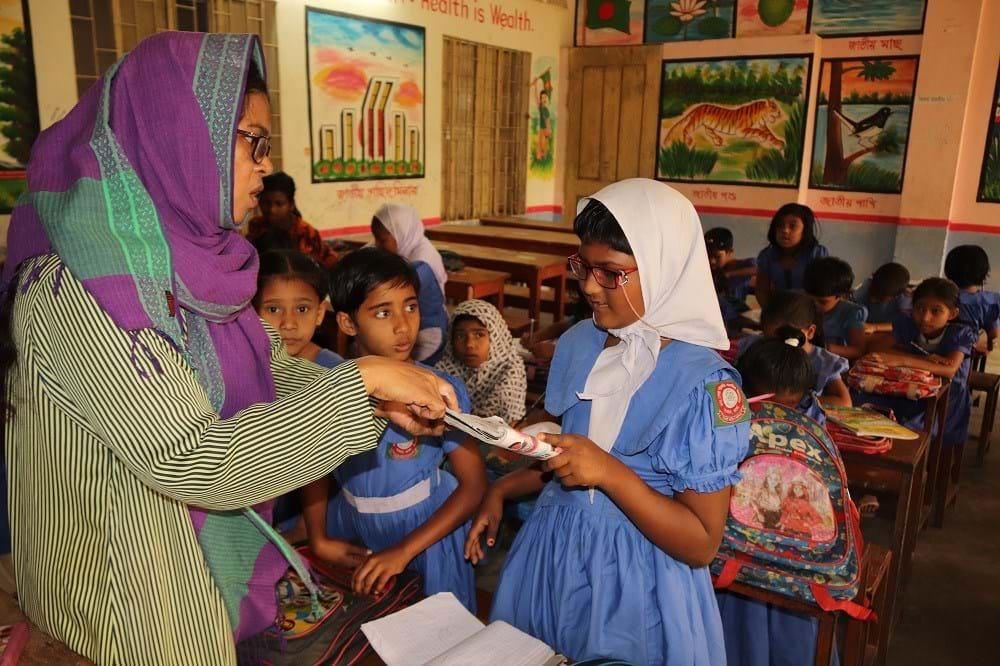 Children in a classrom stading with their teacher. A young girl has a book in her hands