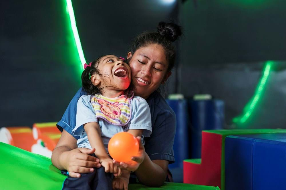 A little girl sitting on the lap of a woman smiling and playing with a ball