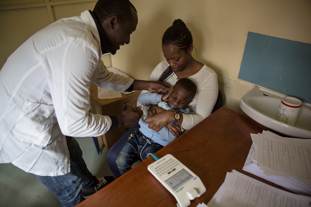 A child sits on her mother's lap taking a hearing test from a doctor