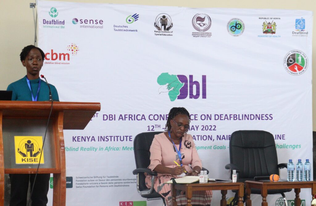 A woman speaking from a lectern and a woman sat at a desk