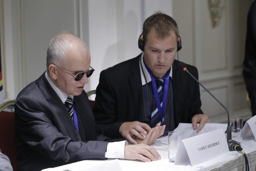 Vasile Adamescu with his interpreter, who is a man both wearing suits sat at a table.