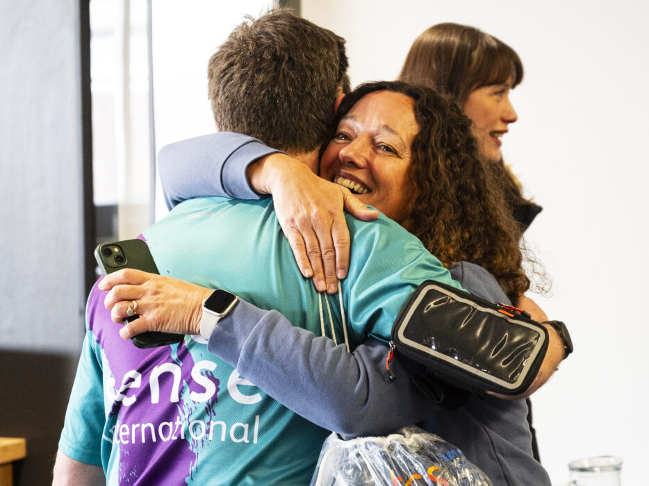 A woman with curly hair hugs a man wearing a Sense International running shirt