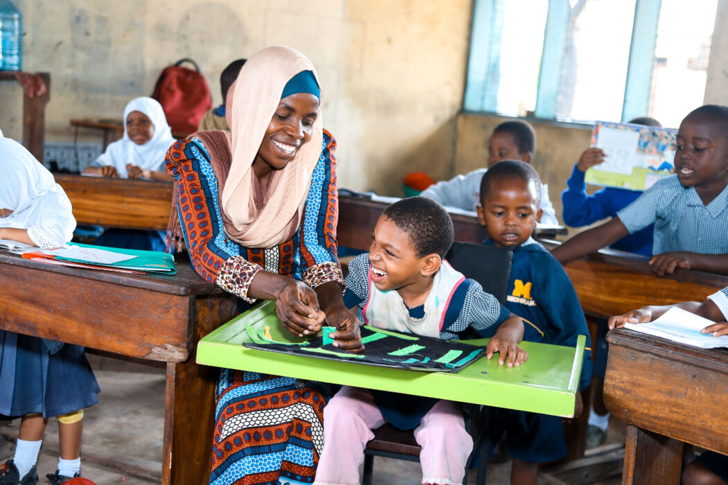 A young school girl sitting at an assistive seating desk laughs with her teacher who smiles at her