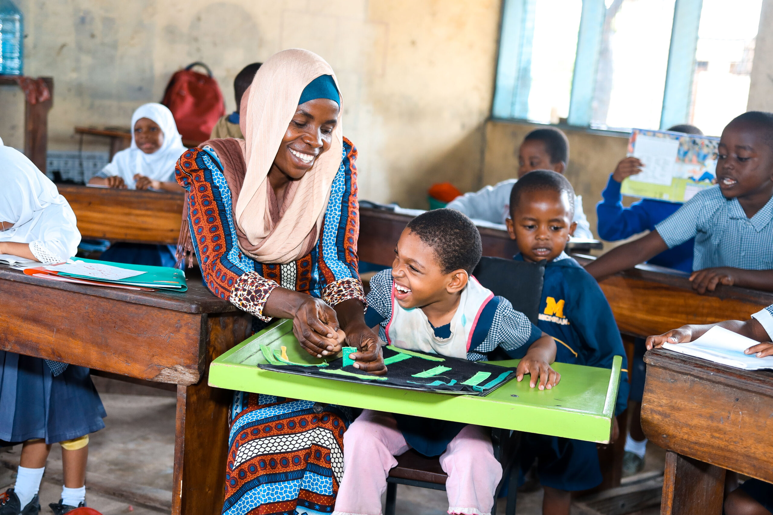 A young school girl sitting at an assistive seating desk laughs with her teacher who smiles at her