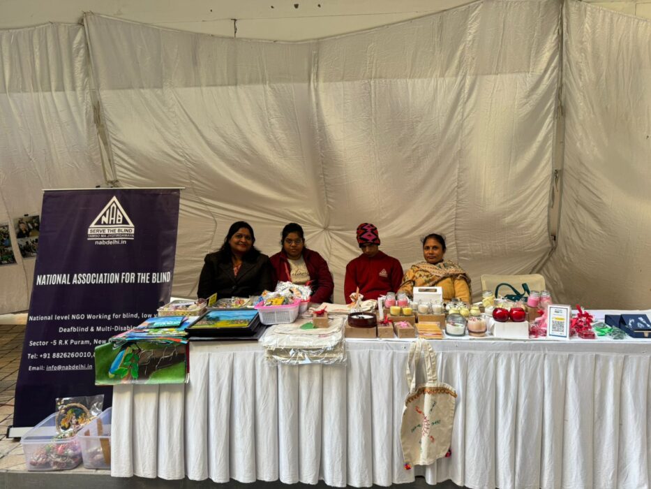 Four people sit behind a long table of food, packages and posters. A banner to their left reads "National Association for the Blind"