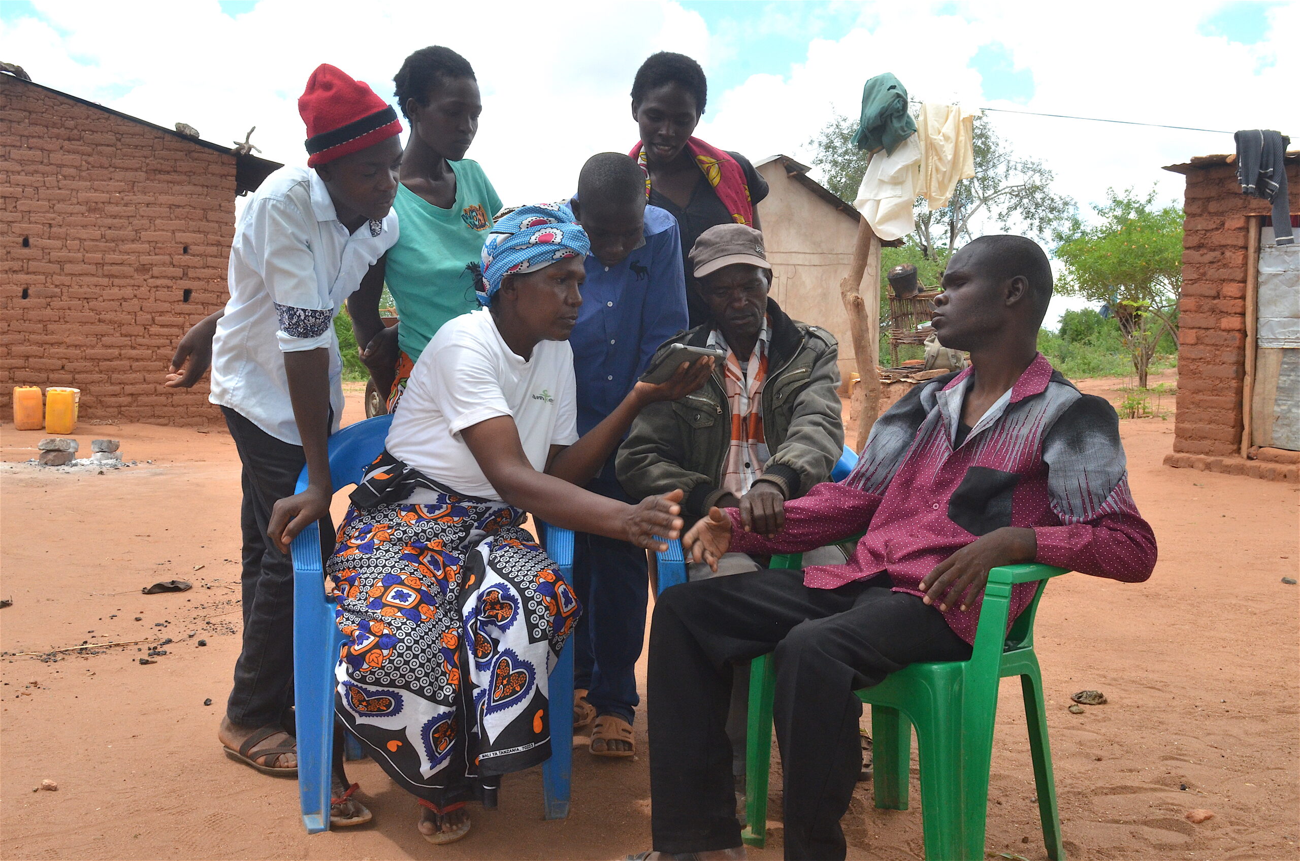Six family members wearing brightly coloured clothing gather around a man sat in a green plastic chair, learning together how to use a digital tablet