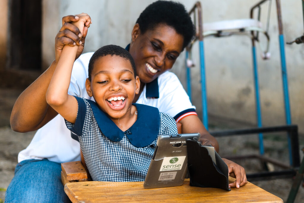 A young child with very short hair wearing a schooldress smiles and laughs with her teaching assistant as they interact with a table with a Sense International sticker on the back