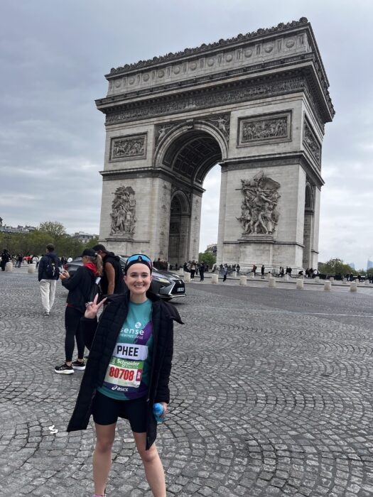 A woman wearing a Sense International running shirt and black coat stands in front of the Arc de Triomphe