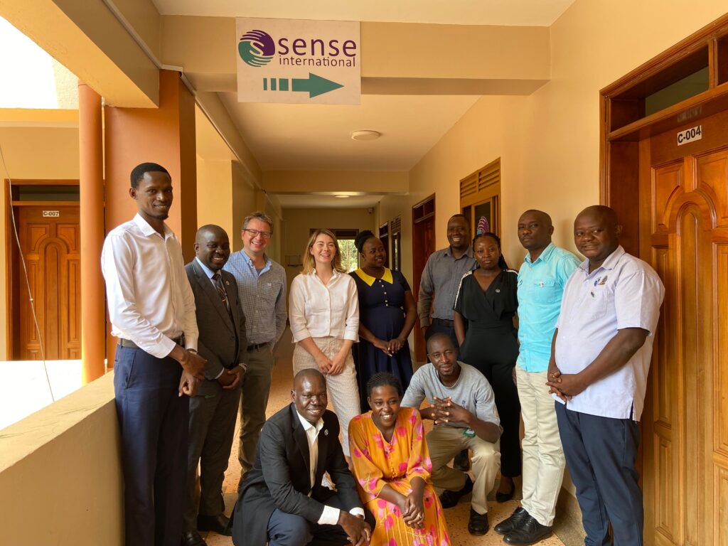 A group of twelve people gathering to smile for the camera under a sign featuring the Sense International logo