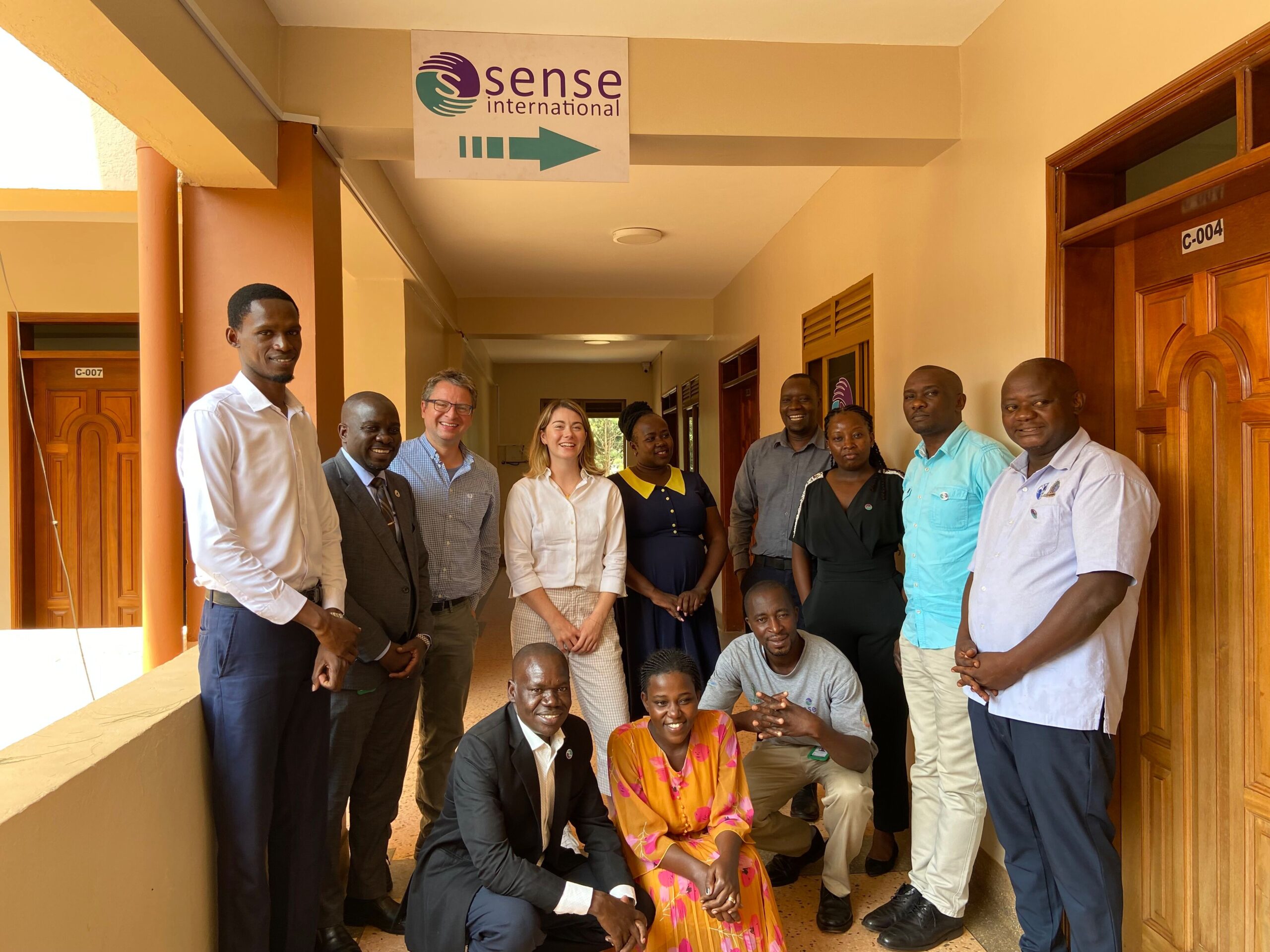 A group of ten people wearing colourful clothing stand together under a sign for Sense International Uganda