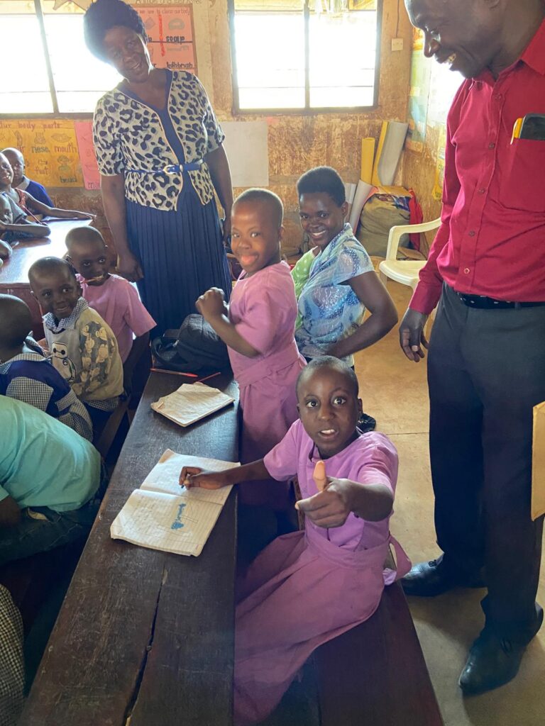 Inside a classroom, two young girls wearing pink small dresses sit at a desk with their teacher and learning support assistant. One gives a thumbs up, the other sticks her tongue out.