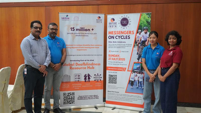 Four people in Sense International India shirts stand in front of a large white banner. Visible text on the banner includes
"15 million + people around the world live with deafblindness"
"Introducing first of its kind Global Deafblindness Resource Hub"

"Messengers On Cycles - Sunday 27 July 2025"