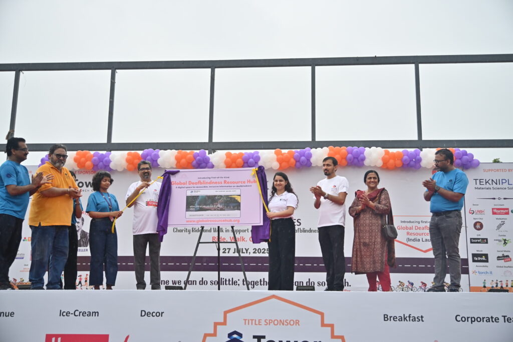 Eight people, many of whom are wearing Sense International India shirts, stand on a large white stage decorated with orange and purple balloons. Two of them are unveiling a large poster with the title 'Global Deafblindness Resource Hub'