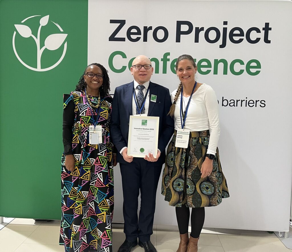 Two staff members from Kenya and one white British woman all wearing colourful clothing stand in front of a green and white sign which reads 'Zero Project Conference'. The man standing in the middle holds a Zero Project Award certificate.
