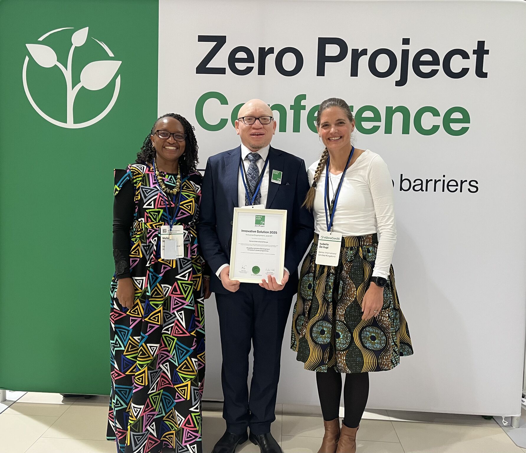 Two staff members from Kenya and one white British woman all wearing colourful clothing stand in front of a green and white sign which reads 'Zero Project Conference'. The man standing in the middle holds a Zero Project Award certificate.
