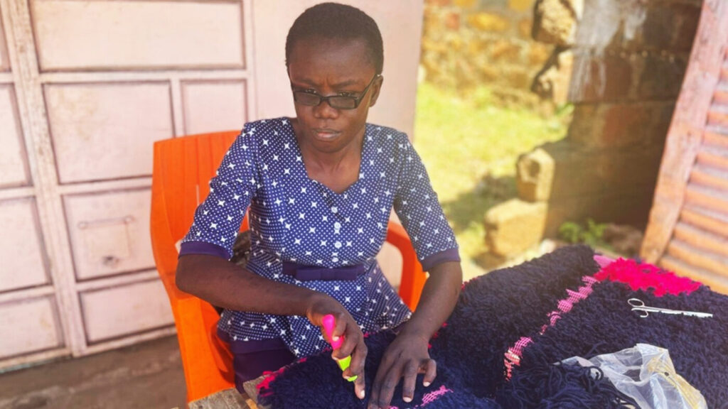 A Kenyan woman wearing a blue dress and dark glasses, crafting at a table