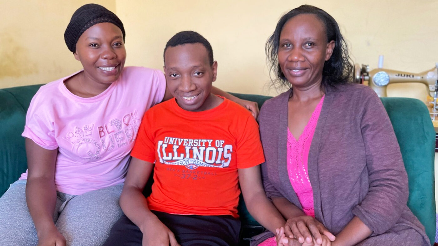 A Kenyan mother and grandmother, both wearing pink, sit either side of a young man wearing a red t-shirt on a sofa.