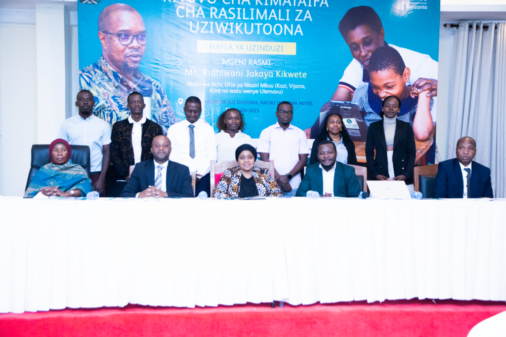 A group of people wearing formal shirts and suits sit at a white cloth table for a press conference, in front of a sign for Sense International Tanzania