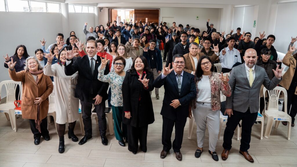 A huge crowd of people wearing formal clothing fill an event space. They are smiling at the camera and making a "horned" hand sign which means "I love you" in American Sign Language
