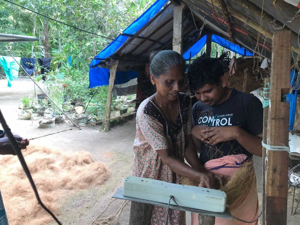 A young Indian man and his elderly mother stand in a yard weaving yarn