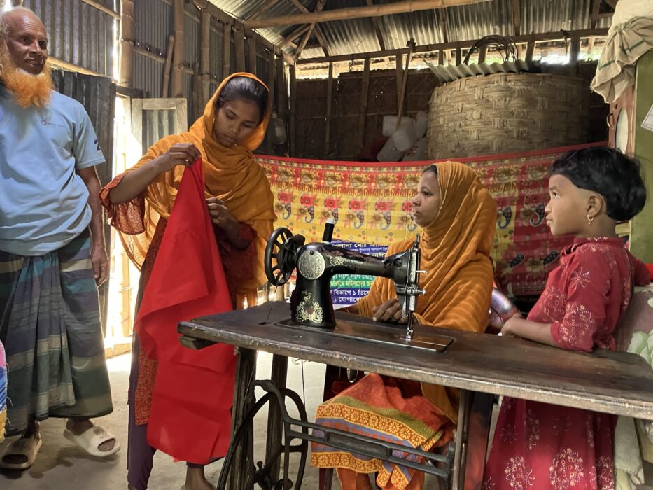 Two young adult Bangladeshi women wearing orange sari sit at a sewing machine, one holding up a vivid red cloth. A young boy and an older man stand by.