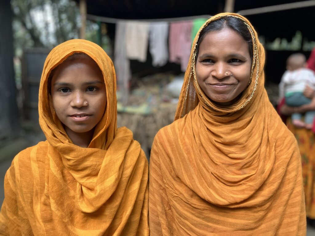 Two young Bangladeshi women wearing bright orange headscarves smile at the camera. 