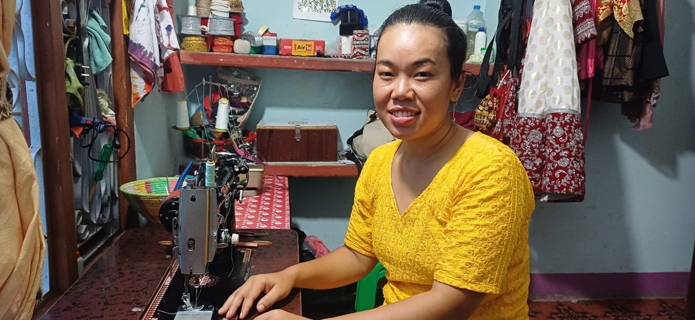 A young Nepali woman in a bright yellow dress smiles, sitting in front of a sewing machine