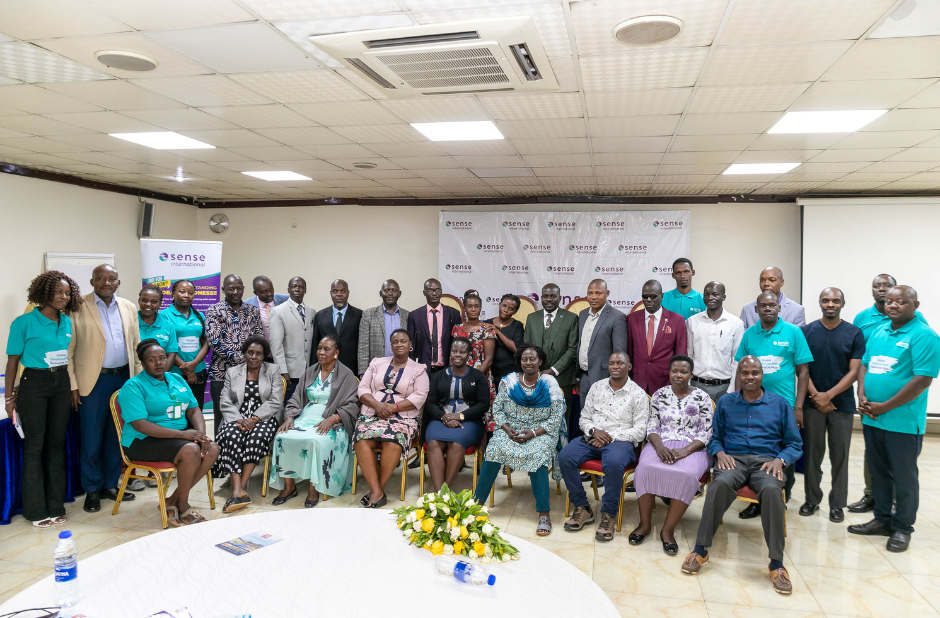 A large crowd of people, many of whom are wearing teal Sense International Uganda t shirts, gather together in position for a photo, in front of a wall featuring the Sense International logo