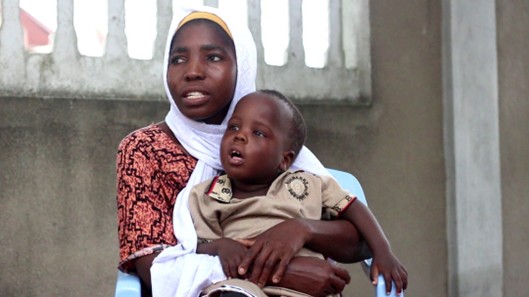 A Tanzanian mother wearing a red dress and white headscarf holds a toddler wearing a beige playsuit.