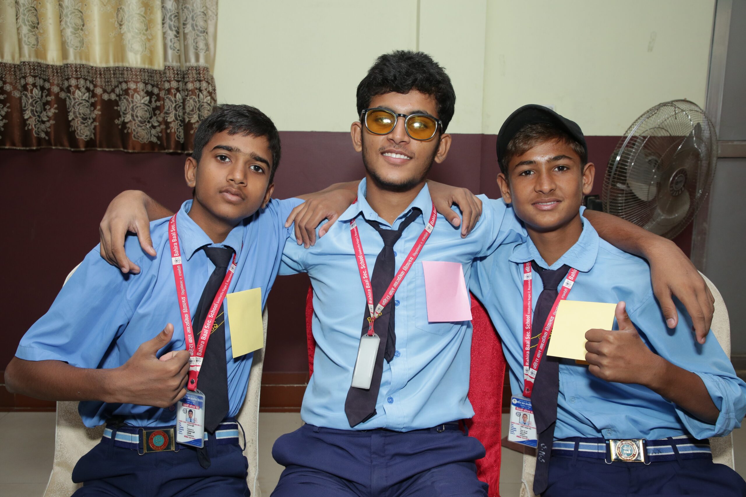 Three Nepali school children wearing blue shirts and ties smile with their arms around one another