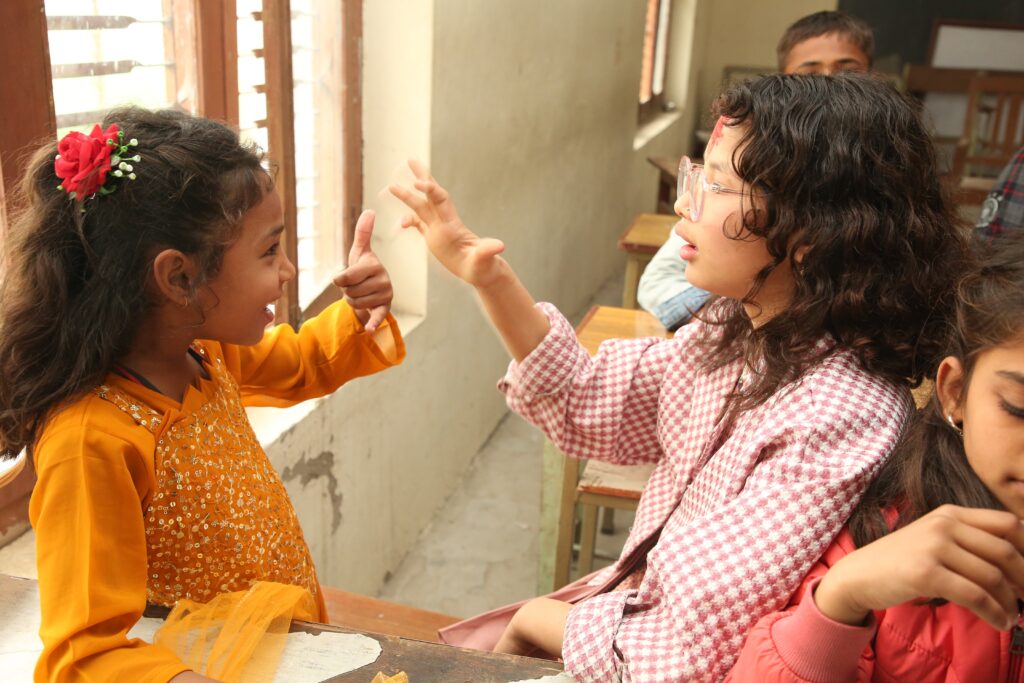 Two young Nepali girls wearing pink and orange dresses communicate in signh language