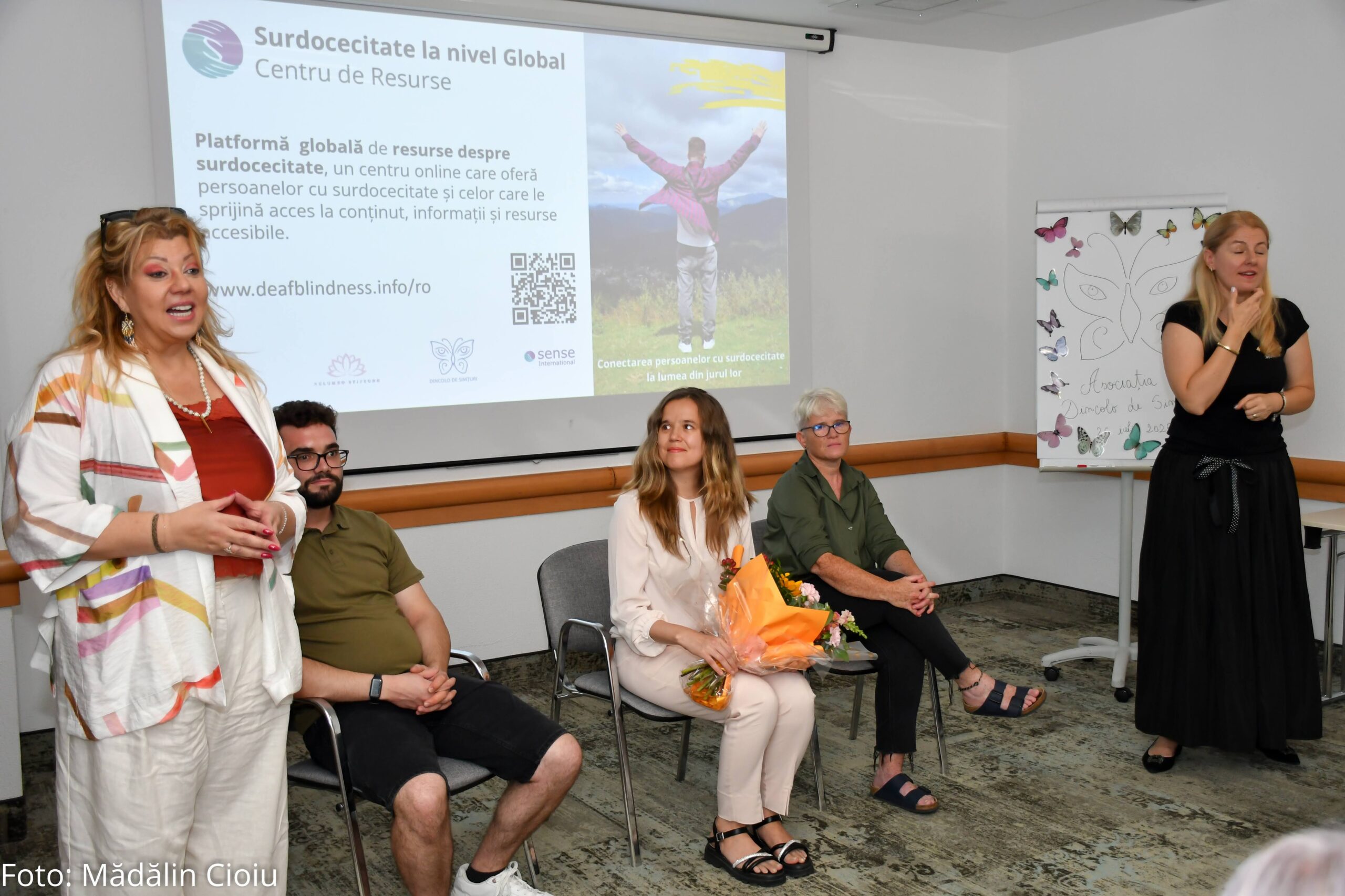 Three young adults sit in chairs in front of a projector screen of text written in Romanian. A woman in a white suit speaks to the crowd whilst another woman in a black dress interprets in sign language.