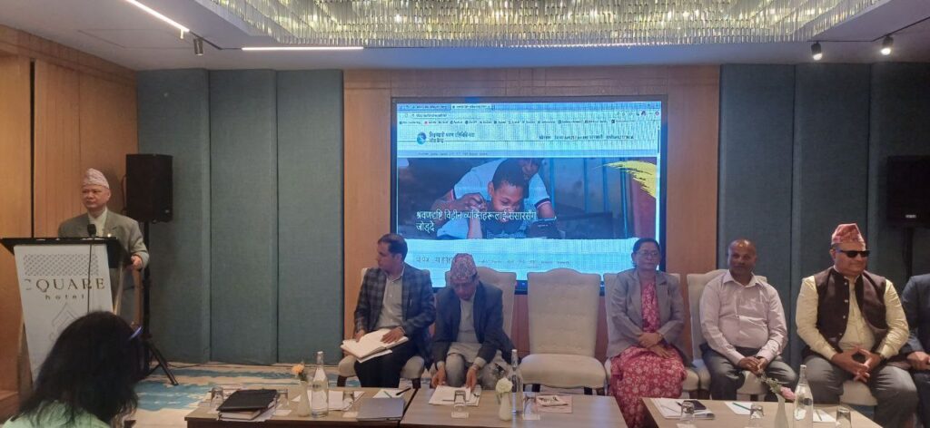 Five people in formal Nepali dress sit in front of a screen featuring images from the Global Deafblindness Resource Hub whilst another man gives a speech at a podium