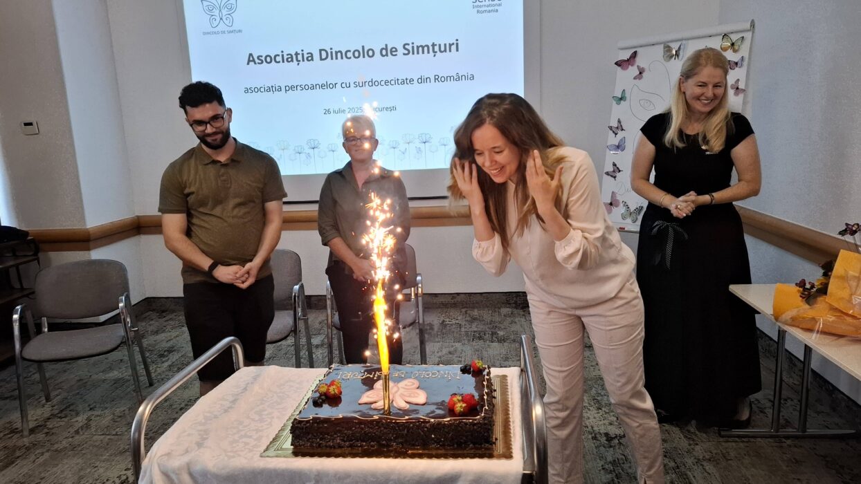 Four young adults standing in a room in front of a projector screen written in Romanian. One women with long hair dressed in beige is blowing out a sparkling candle on a large chocolate cake.