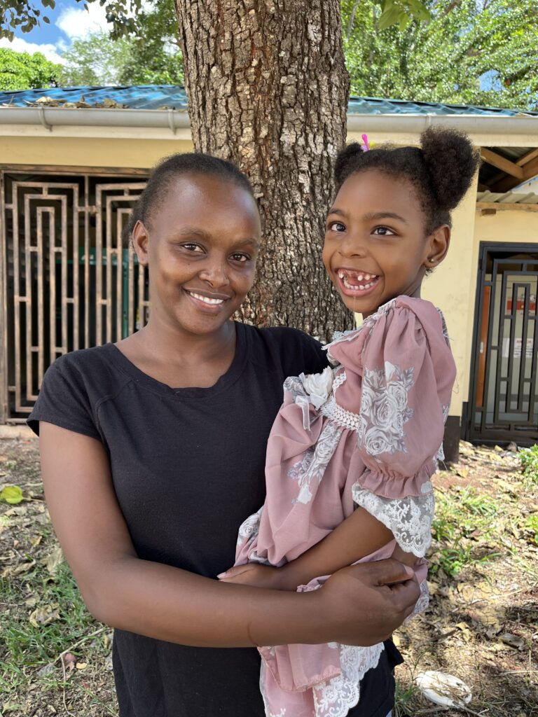 A young Kenyan mother with very short hair holds her toddler daughter who wears a pink dress and bunches