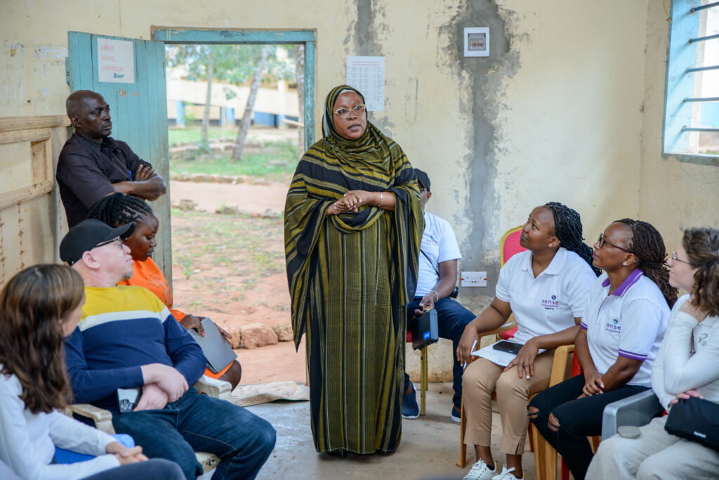 A Kenyan mother wearing a full body headscarf and gown stands at the centre of a room of doctors as she tells her story