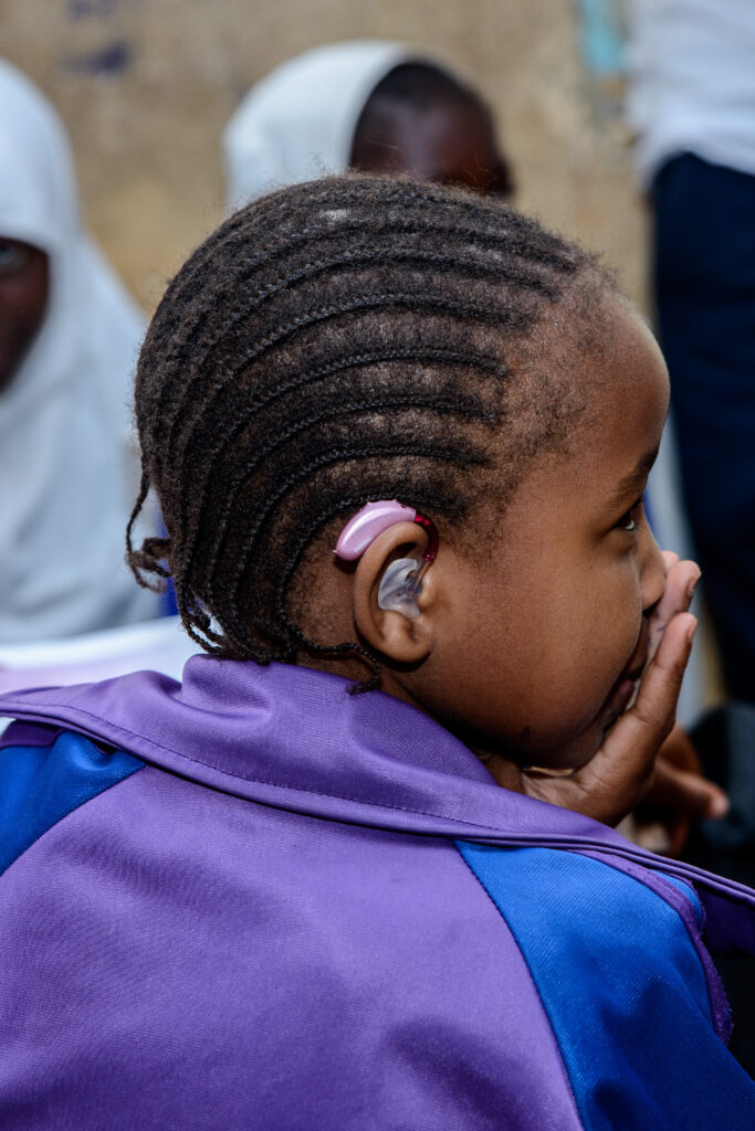 A young Kenyan girl wearung a purple tracksuit faces away from the camera, showing a pink hearing aid attached to the back of her ear