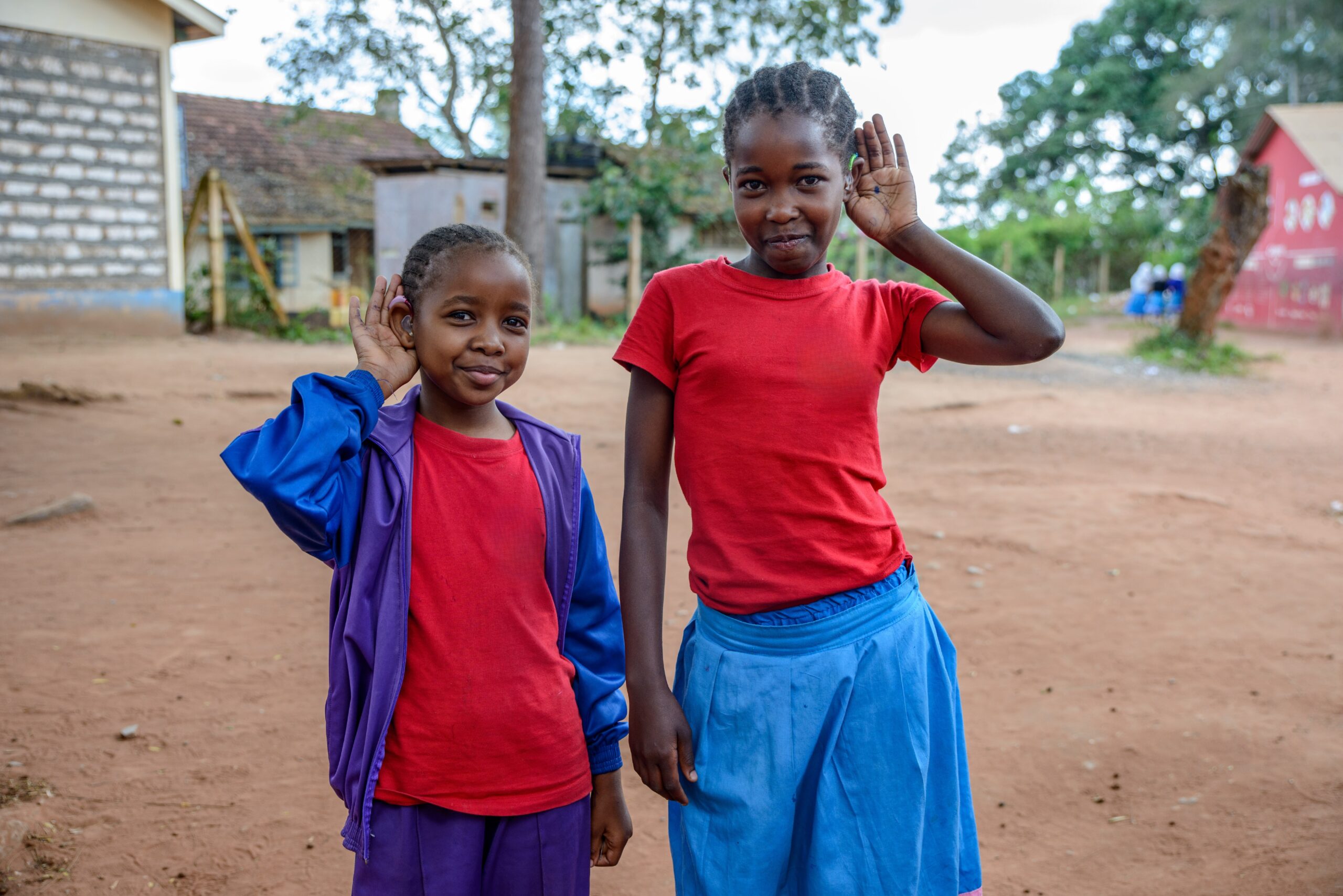 Two young Kenyan girls wearing bright red t-shirts cups their heads to their ears, showing off their hearing aids.