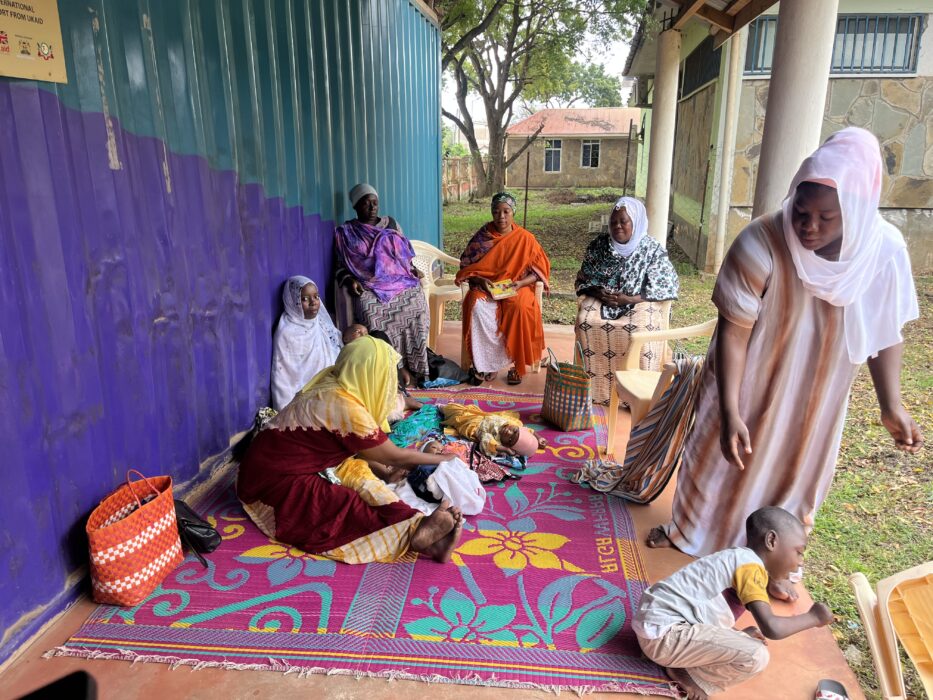 A group of Kenyan mothers wearing brightly coloured clothing sit on a colourful mat with their babies