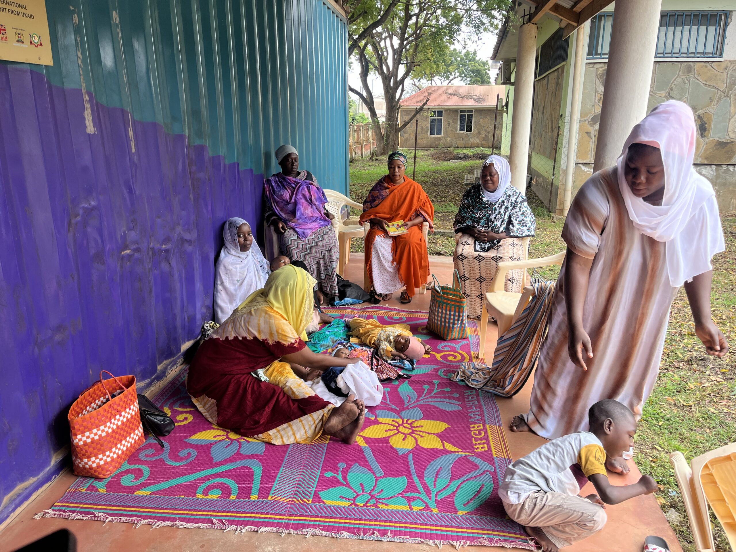 A group of Kenyan mothers wearing brightly coloured clothing sit on a colourful mat with their babies