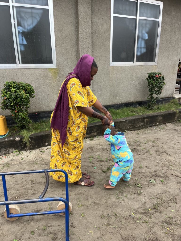 A woman wearing a bright yellow dress and purple headscarf holds the hand of a toddler wearing bright clue clothes to help him stand