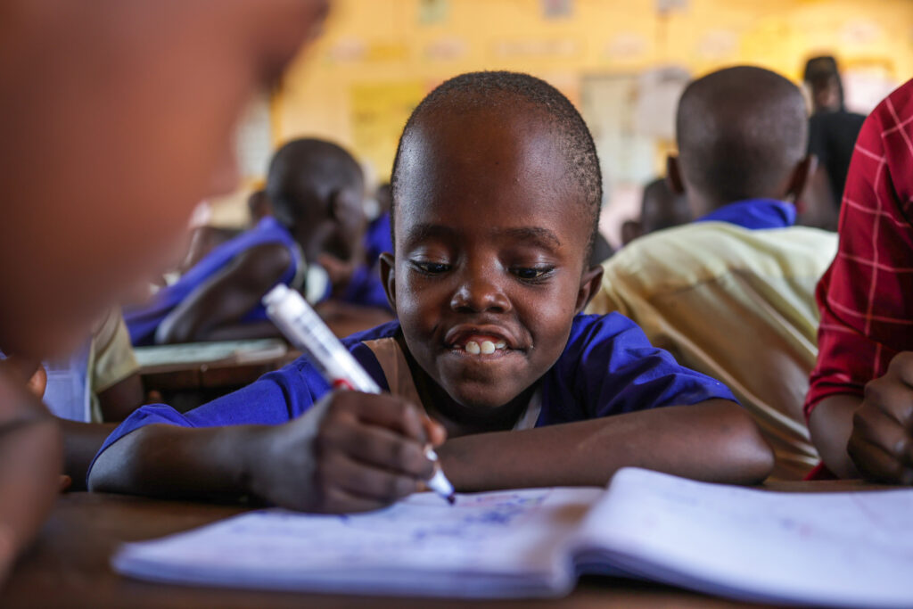 A young Ugandan child with a very short hair wearing a blue school uniform smiles as she writes in a notebook
