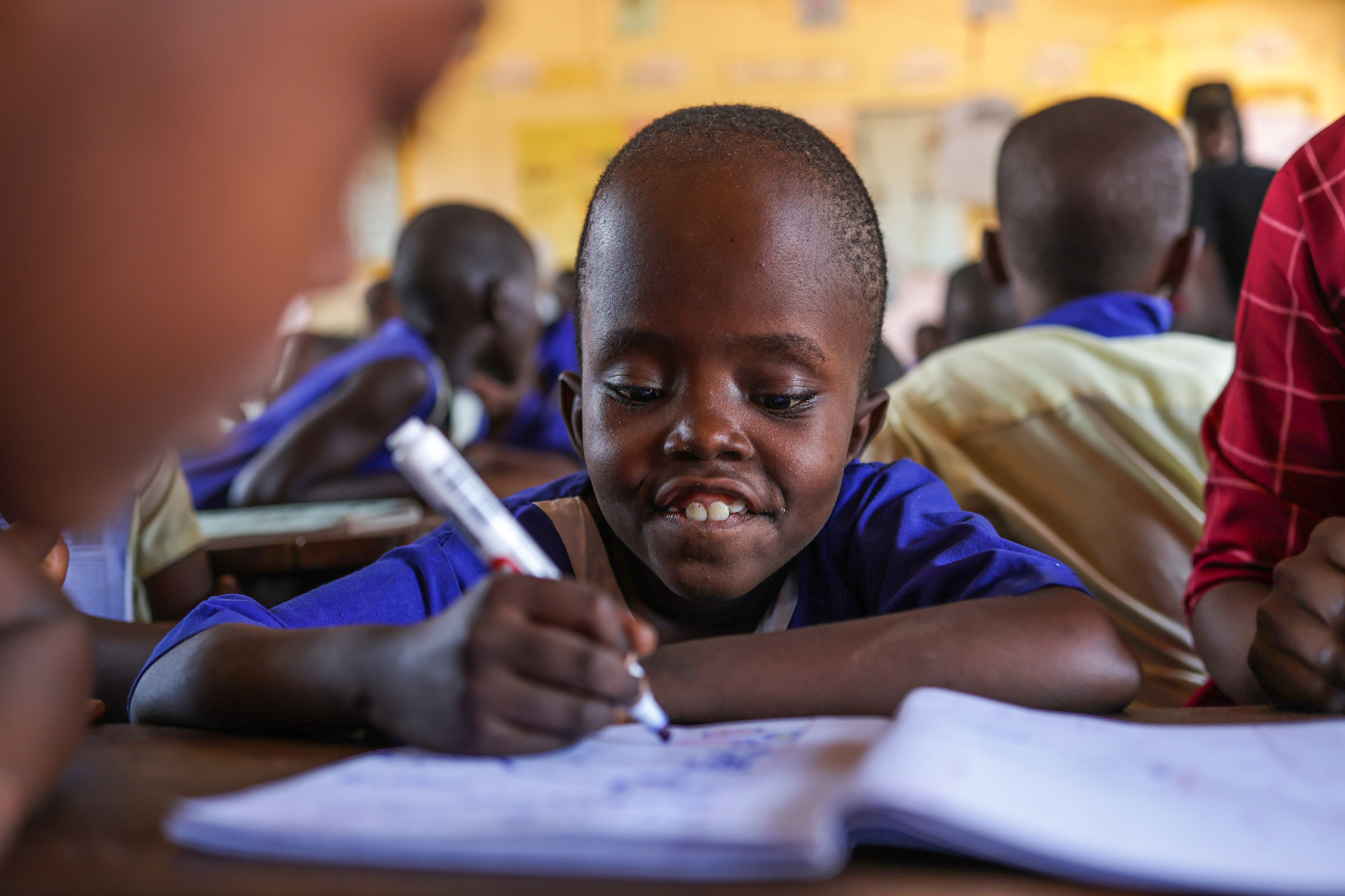 A young Ugandan child with a very short hair wearing a blue school uniform smiles as she writes in a notebook