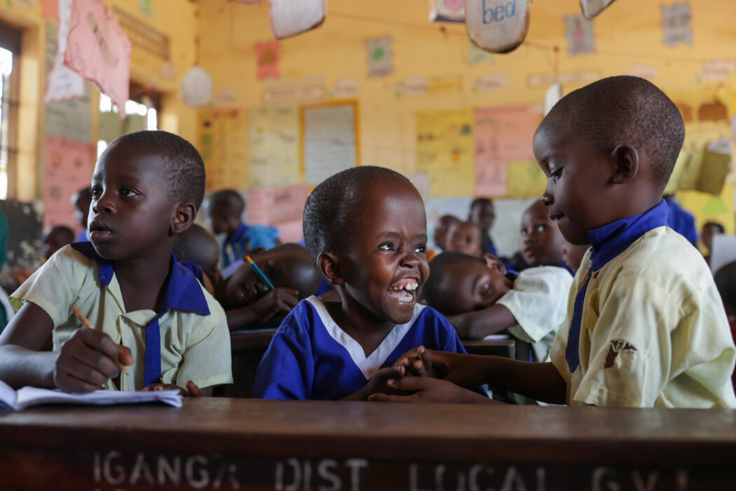 In an orange classroom, a young Ugandan child with a very short hair wearing a blue school uniform laughs excitedly with her classmates