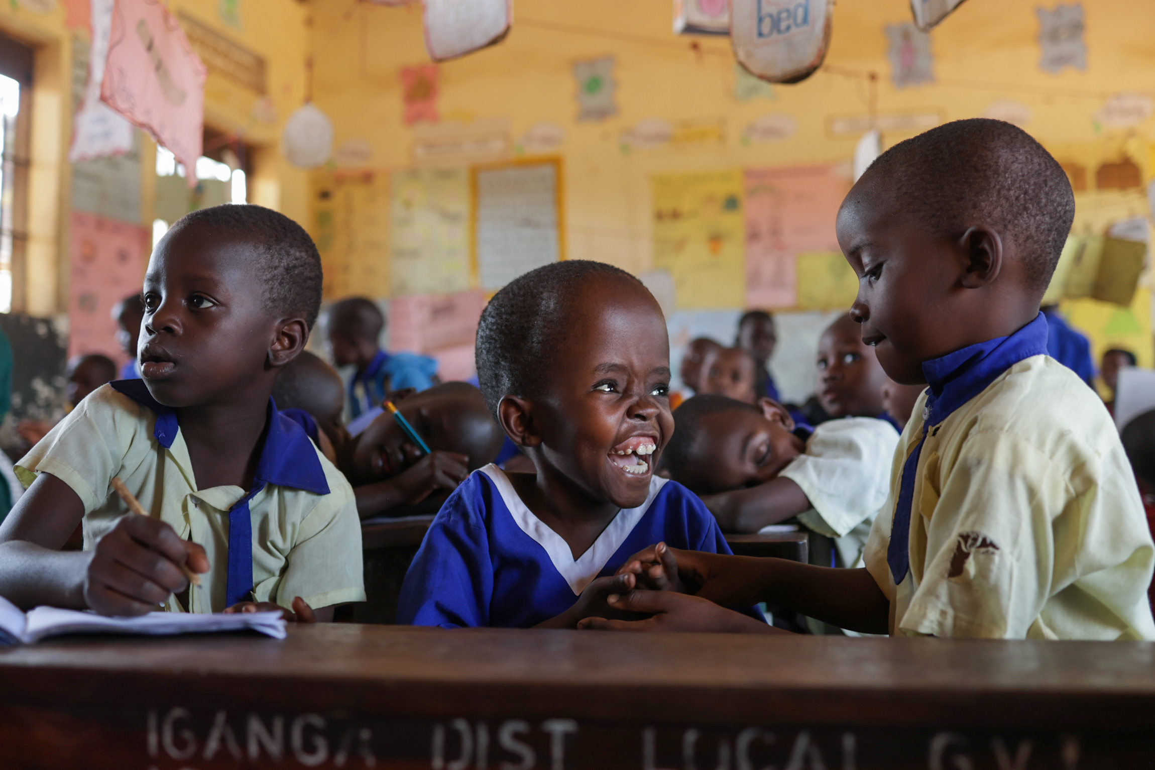 In an orange classroom, a young Ugandan child with a very short hair wearing a blue school uniform laughs excitedly with her classmates
