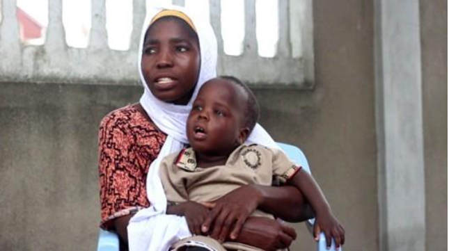 A young Tanzanian woman wearing a red dress and white headscarf looks to the left, holding her toddler who wears a beige playsuit.