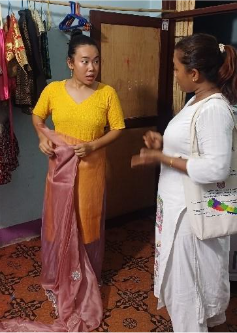 A young adult Nepali woman wearing a bright yellow dress holds a pink dress, looking at a woman dressed in white