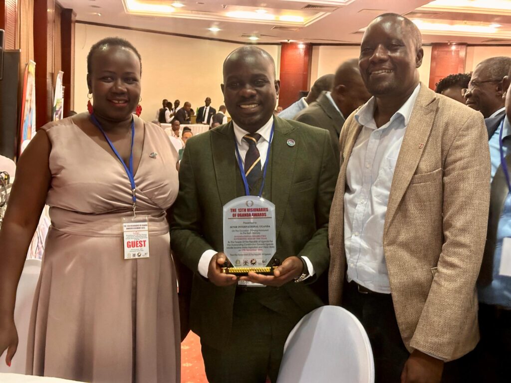 A woman wearing a pink dress and two men wearing black and beige suits hold an award reading 'The 13th Visionaries of Uganda Awards 2025'
