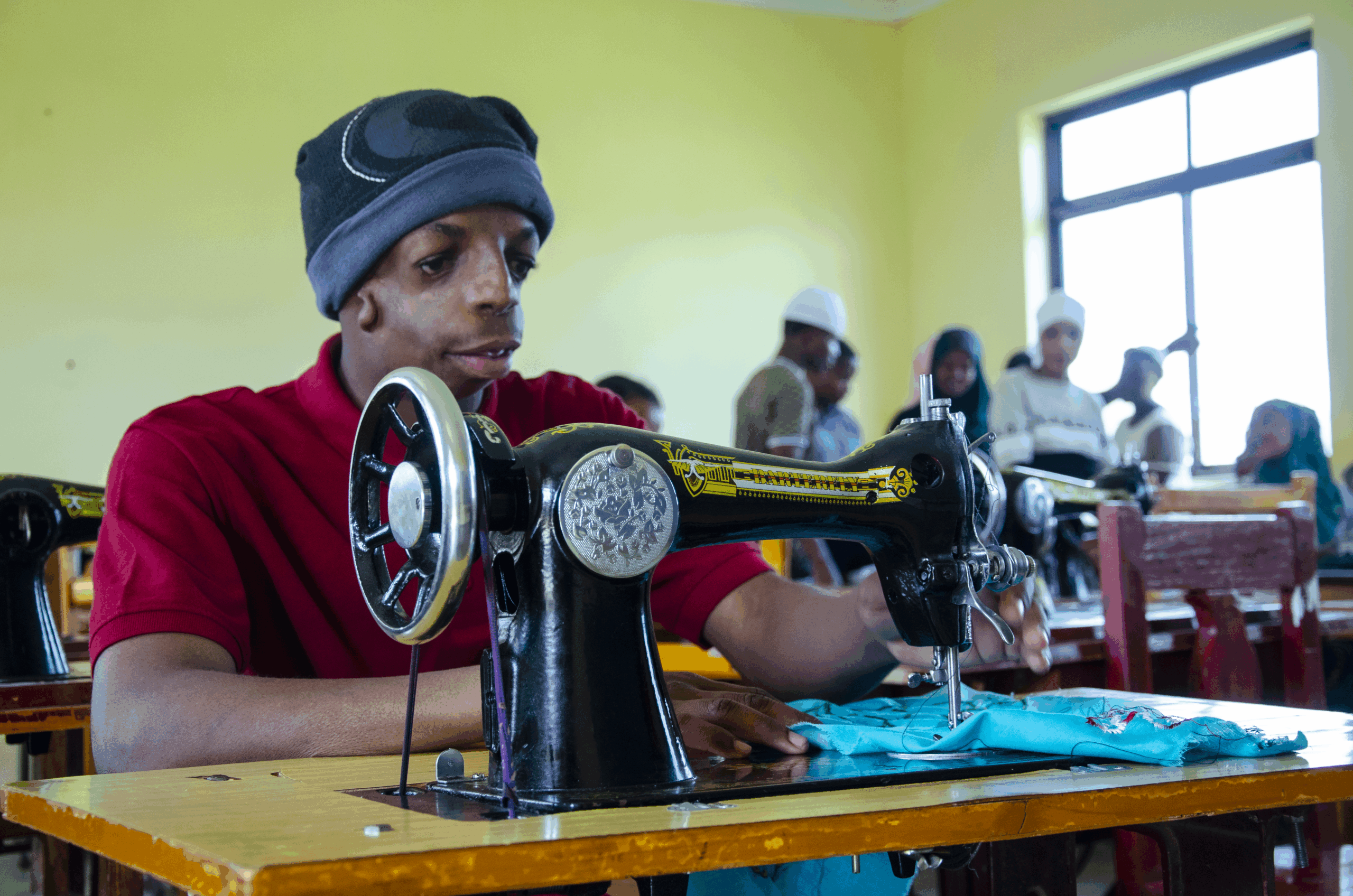 A young Tanzanian man wearing a blue wooly hat and red shirt using a sewing machine