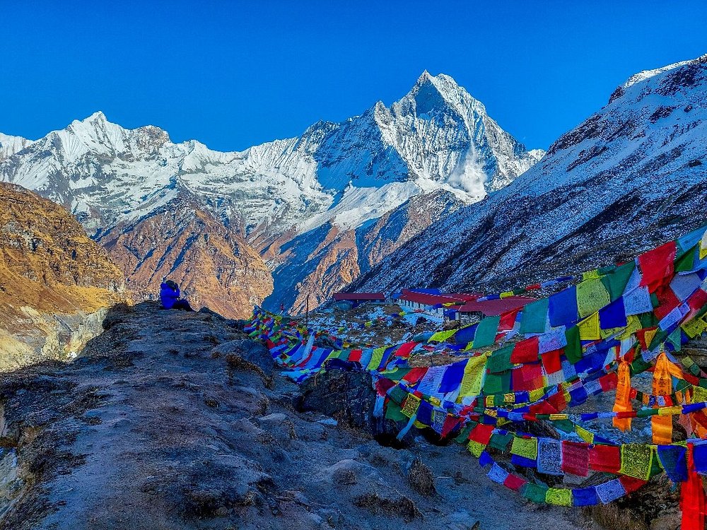 The snowy peaks of Annapurna are lit by the sun in the background, with a man in the distance sitting on the edge of the mountain. Swathes of colourful flags are visible in the background.